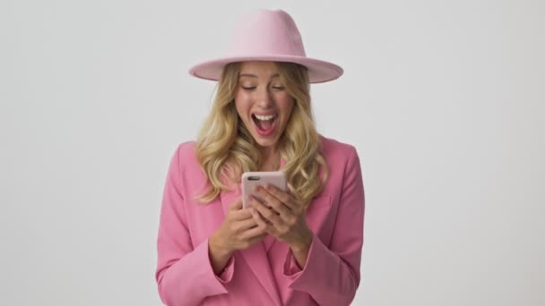 ピンクのジャケットと帽子の魅力的な若いブロンドの女性は非常に幸せになり 灰色の背景の上にスマートフォンを使用しながら 手で勝者のジェスチャーを作ります — ストック動画