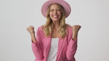 Pembe ceket ve şapka neşeli genç sarışın kadın iki eliyle kazanan jest yapma ve gri arka plan izole üzerinde kameraya bakarken çığlık
