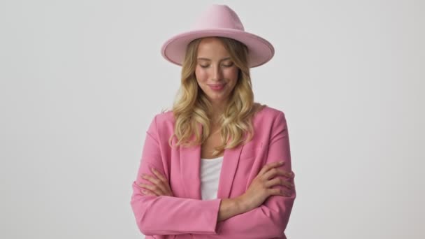 ピンクのジャケットと帽子をかぶったかわいい若いブロンドの女性は 灰色の背景の上にカメラを見ながら笑顔で腕を組んだ — ストック動画