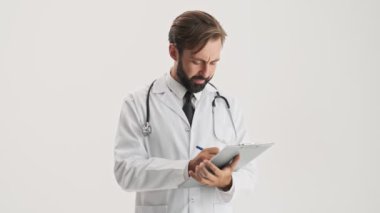 Stetoskop ile beyaz profesyonel ceket üzgün genç sakallı adam doktor tıbbi geçmişi bakarak ve gri arka plan izole üzerinde olumsuz başını sallayarak