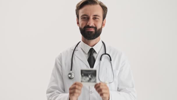 聴診器の笑顔と灰色の背景の上に赤ちゃんの超音波スキャンを示す白いプロのコートで幸せな若いひげの男の医者 — ストック動画