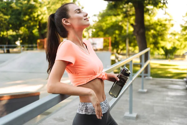 Imagen de linda chica de fitness en ropa deportiva sonriendo y sosteniendo la botella de agua en el campo de deportes — Foto de Stock