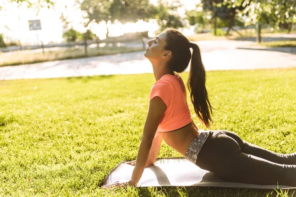 Immagine di una bella giovane donna che fa esercizi di fitness sul tappetino yoga nel parco verde — Foto Stock