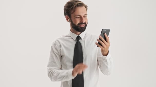 灰色の背景の上にスマートフォン上のビデオ通話を持つワイヤレスイヤホンを持つ魅力的な若いひげのビジネスマン — ストック動画