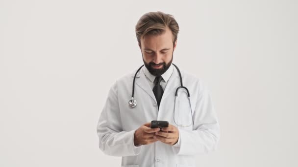 年轻英俊的留着胡子的医生 身穿白色职业装 手持听诊器 用智能手机 面带微笑 在灰色的背景下孤立地望着相机 — 图库视频影像