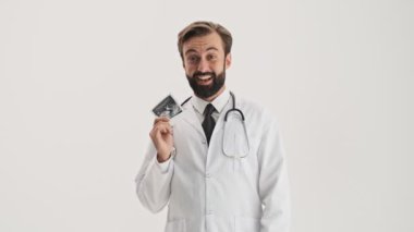 Steteskoplu beyaz önlüklü heyecanlı genç sakallı doktor gri arka planda bebeğinin ultrason taramasını gösteriyor.