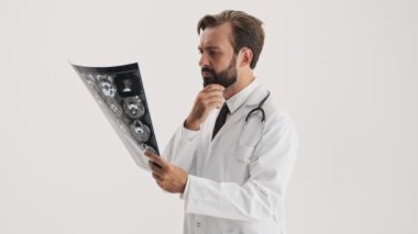 Steteskoplu beyaz önlüklü, ciddi, sakallı, sakallı, sakallı ve sakallı bir doktor. Gri arka plandaki röntgen görüntüsüne dikkatlice bakıyor.