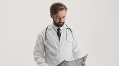 Steteskoplu, beyaz profesyonel ceketli çekici genç sakallı doktor. Gri arka plandaki röntgen görüntüsüne dikkatle bakarken gülümsüyor ve baş parmağını kaldırıyor.