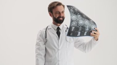 Stetoskop gülümseyerek ve gri arka plan izole üzerinde iki x-ray görüntüleri dikkatle bakarak beyaz profesyonel ceket Mutlu genç sakallı adam doktor