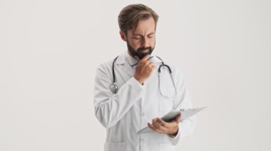 Disappointed thoughtful young bearded man doctor in white professional coat with stethoscope touching his beard and nose bridge while working with documents attentively over gray background isolated