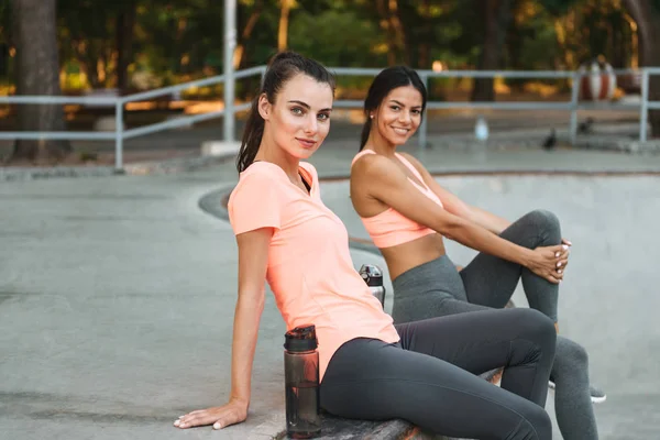 Imagem de mulheres atléticas sorridentes sentadas com garrafas de água no chão de esportes de concreto — Fotografia de Stock