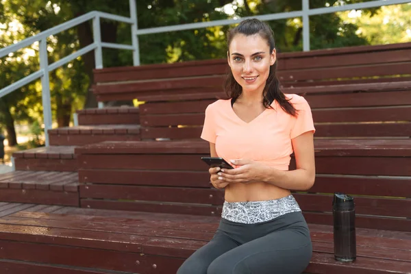 Immagine di una bella ragazza in abbigliamento sportivo che tiene il cellulare mentre è seduta sulla panchina nel parco — Foto Stock