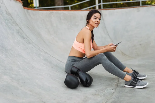 Bild av fitness Young Girl Holding mobil medan du sitter på betonggolv med boxningshandskar — Stockfoto