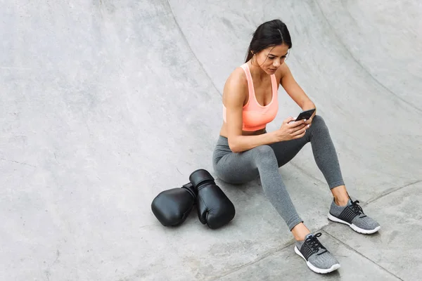 Imagen de una joven caucásica sosteniendo el teléfono celular mientras está sentada en el piso de concreto con guantes de boxeo — Foto de Stock