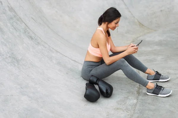 이미지 의 갈색 머리 어린 소녀 들고 휴대 전화 동안 앉아 있는 콘크리트 바닥에 와 권투 장갑 — 스톡 사진