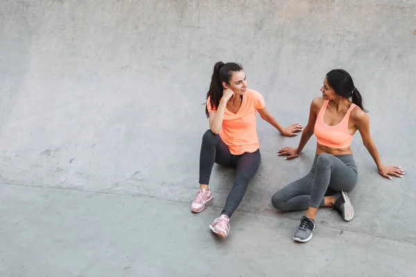 Зображення привабливих фітнес-жінки в спортивному одязі, що розмовляють разом, сидячи на конкретному спортивному майданчику — стокове фото