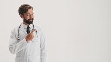 Smiling young bearded man doctor in white professional coat with stethoscope pointing with finger to the side and making thumb up gesture while looking at the camera over gray background isolated
