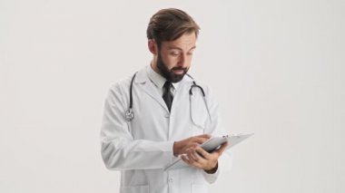 Serious young bearded man doctor in white professional coat with stethoscope looking at the medical history and shaking his head negatively over gray background isolated