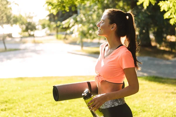 Immagine della giovane gioiosa ragazza che tiene in mano la bottiglia d'acqua mentre cammina con il tappetino da fitness nel parco — Foto Stock