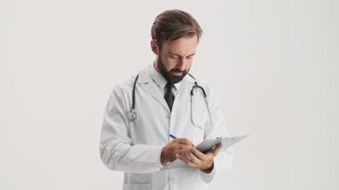 Stetoskop lu beyaz profesyonel paltolu konsantre genç sakallı adam doktor, gri arka plan izole üzerinde özenle belgelerle çalışıyor