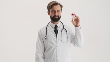 Stetoskop ile beyaz profesyonel ceket çekici genç sakallı adam doktor hapları ile bir şişe atma ve gri arka plan izole üzerinde kameraya bakarken parmak sallayarak hiçbir işaret gösteren
