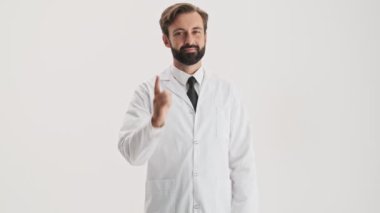 Smiling young bearded man doctor in white professional coat winking and pointing with finger to the camera over gray background isolated