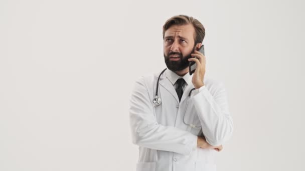 灰色の背景の上にスマートフォン上で非常に迷惑な電話を持つ聴診器を持つ白いプロのコートで不愉快な若いひげの男の医者 — ストック動画