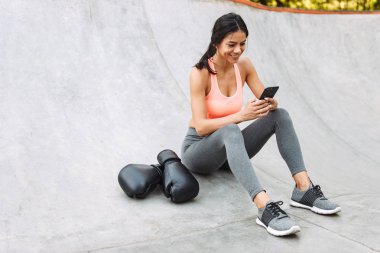 Image of pretty young girl holding cellphone while sitting on concrete floor with boxing gloves clipart