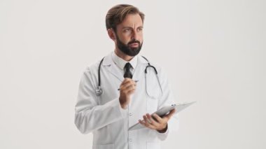 Concentrated young bearded man doctor in white professional coat with stethoscope working with documents attentively and making notes over gray background isolated