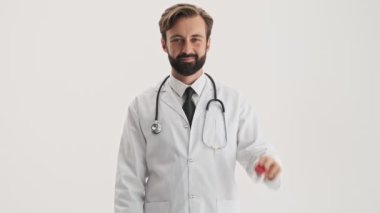 Handsome young bearded man doctor in white professional coat with stethoscope smiling and pointing with finger at the bottle with pills while looking at the camera over gray background isolated