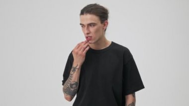 Pensive young tattooed brunette man in black t-shirt thinking about something and touching his face over gray background isolated