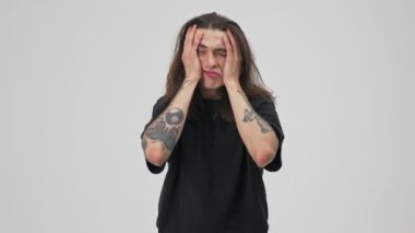 Displeased crazy young tattooed brunette man with long hair in black t-shirt feeling nervous and depressed while rubbing his face with hands over gray background isolated