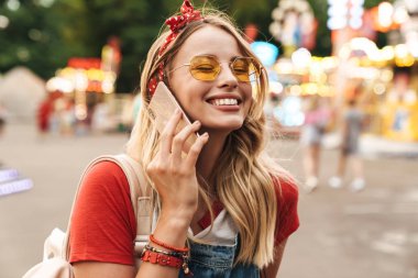Gülümseyen memnun genç neşeli sarışın kadın eğlence parkı cep telefonu ile konuşurken.