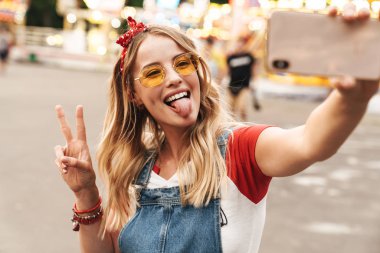Eğlence parkında gülümseyen mutlu neşeli genç sarışın kadın cep telefonu ile selfie çekmek.