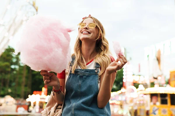Зображення щасливої блондинки, що їсть солодку бавовняну цукерку під час прогулянки — стокове фото