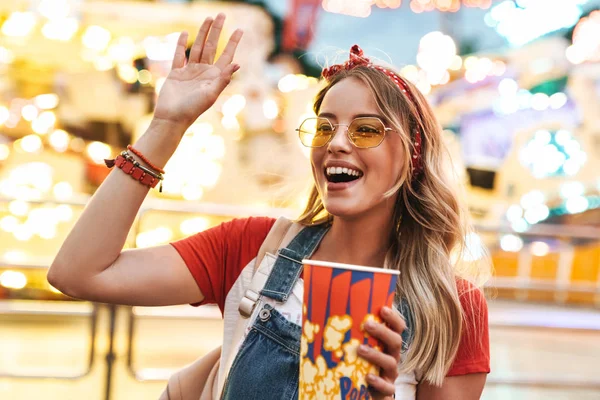 Obraz veselé blondýny mával a držel popcorn, zatímco — Stock fotografie