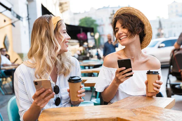 Glada unga nöjda optimistiska flickor vänner sitter utomhus i Café dricka kaffe med mobiltelefoner. — Stockfoto