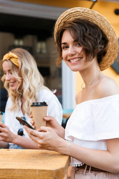 Alegre joven complacido chicas optimistas amigos sentados al aire libre en la cafetería beber café con teléfonos móviles . —  Fotos de Stock