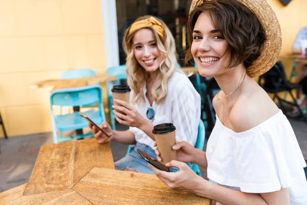 Joyeux jeunes filles heureuses optimistes amis assis à l'extérieur dans le café à boire du café à l'aide de téléphones mobiles . — Photo