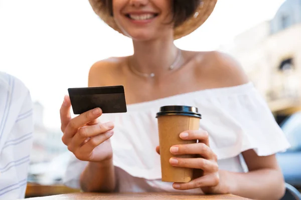 Cheerful young optimistic girl sitting outdoors in cafe drinking coffee holding credit card. — Stock Photo, Image
