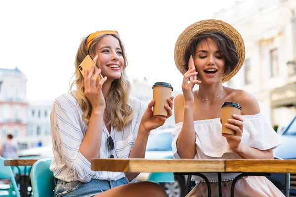 Jóvenes optimistas amigas sentadas al aire libre en la cafetería tomando café hablando por teléfonos móviles . — Foto de Stock