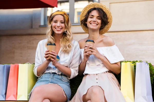 Optimista feliz jóvenes amigas sentadas al aire libre beber café hablando entre sí cerca de bolsas de compras . —  Fotos de Stock