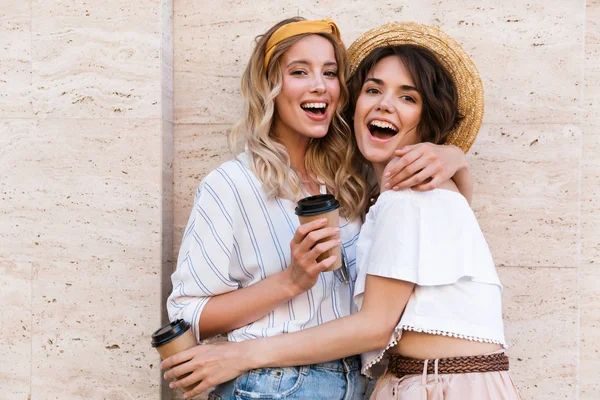 Glad leende positiva unga flickor vänner poserar utomhus dricka kaffe kramas. — Stockfoto