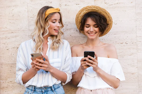 Sonriendo feliz feliz positivo jóvenes amigas al aire libre utilizando teléfonos móviles de chat . —  Fotos de Stock