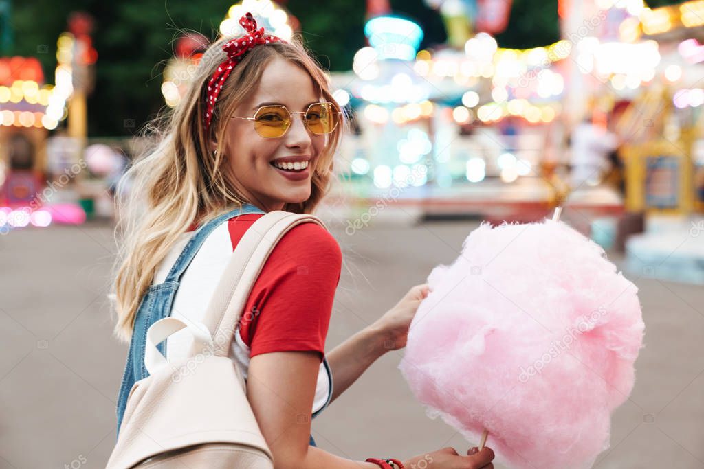 Image of smiling young woman eating sweet cotton candy while wal