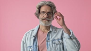 Handsome elderly stylish bearded man with gray hair in denim jacket taking off his eyeglasses and winking while looking at the camera over pink background isolated