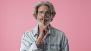 Serious elderly stylish bearded man with gray hair in denim jacket showing silence gesture with finger while looking at the camera over pink background isolated