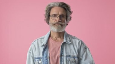 Upset elderly stylish bearded man with gray hair in denim jacket feeling tired and wiping the sweat of his forehead while looking at the camera over pink background isolated