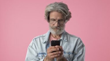 Surprised elderly stylish bearded man with gray hair in denim jacket becoming happy and smiling while using smartphone over pink background isolated