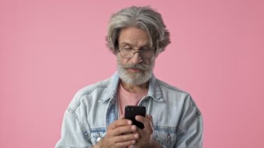 Cheerful elderly stylish bearded man with gray hair in denim jacket screaming and making winner gesture with hand while using smartphone over pink background isolated